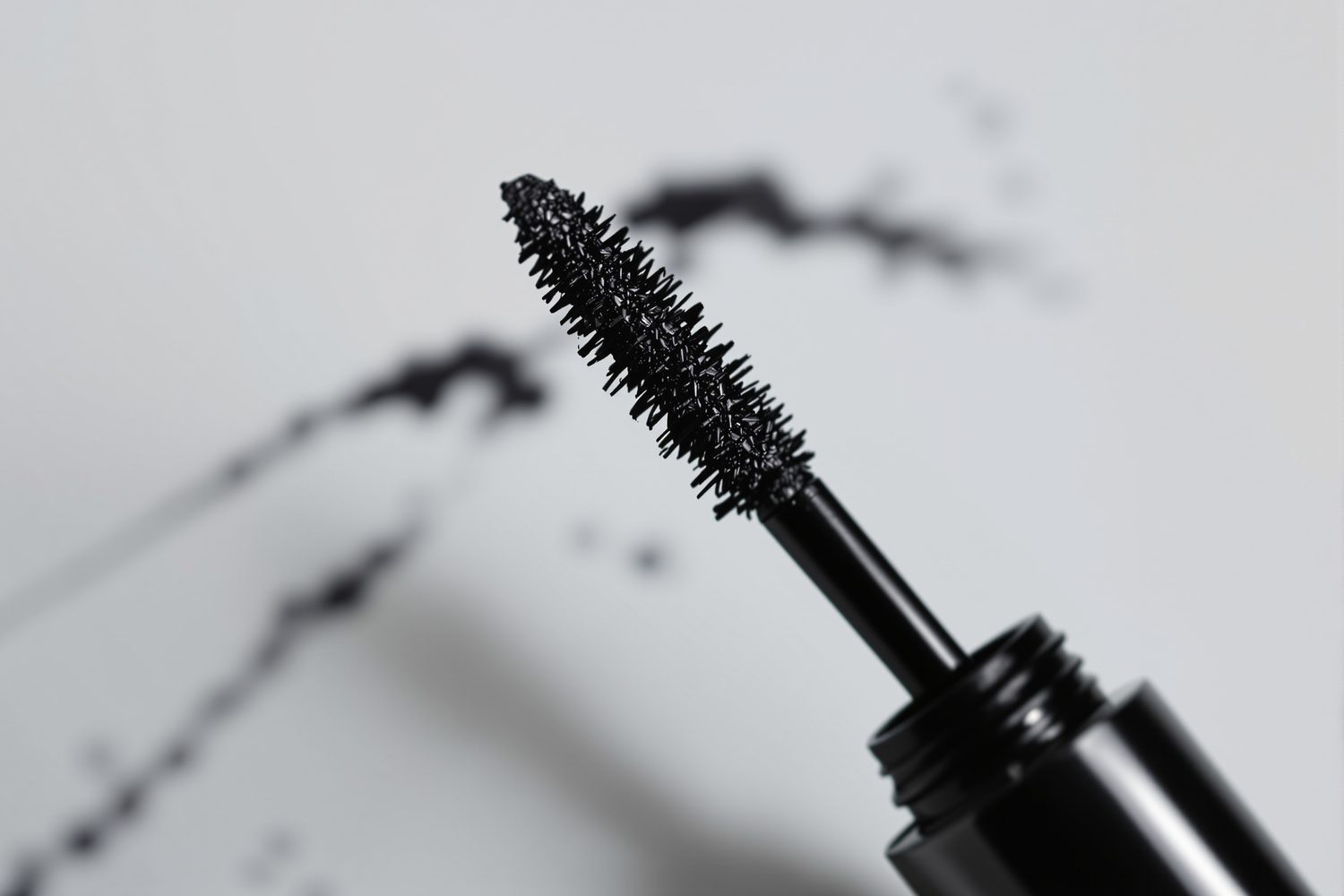 Close-up of a black waterproof mascara wand lifted from its tube, with mascara strokes on a pale grey surface behind it