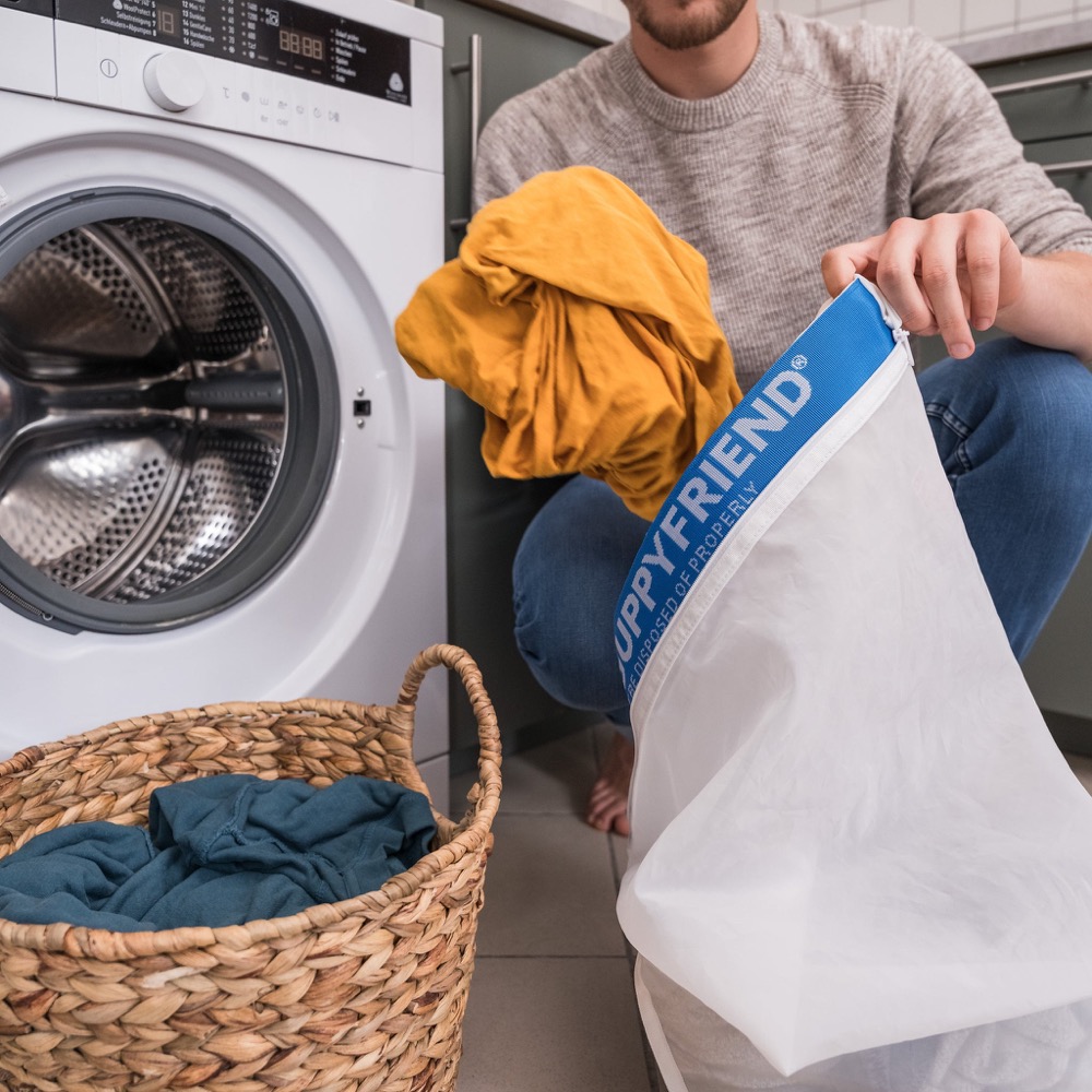 Guppyfriend washing bag — white mesh laundry bag being loaded with synthetic clothing beside a washing machine