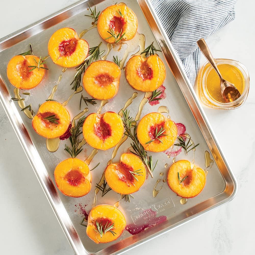 Nordic Ware Naturals aluminum half sheet pan holding roasted peach halves with rosemary and honey, photographed from above on a white counter