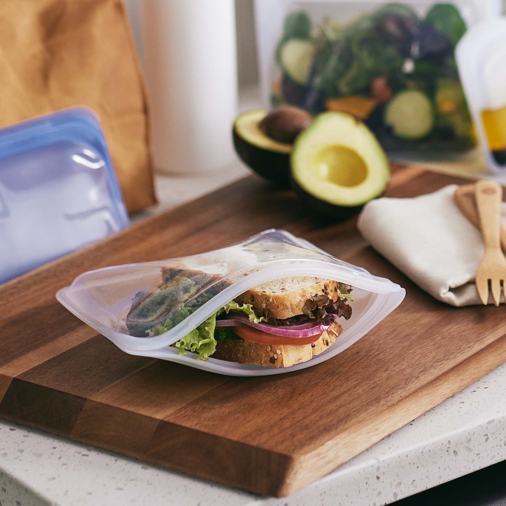 Stasher reusable silicone bag holding a sandwich on a wooden board, with a second bag and avocado nearby